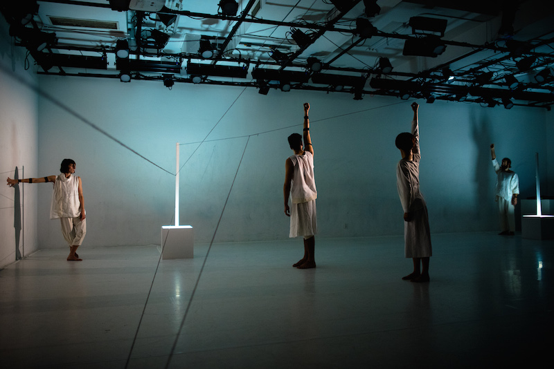 Three dancers stand with the right arms aboce their heads. Light emits from two long sticks that are affixed in cubes. Another dancer traces her hand along the wall.